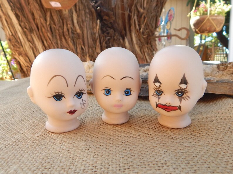 May include: Three porcelain doll heads with painted faces. The doll heads have blue eyes, painted eyebrows, and red lips. One doll head has black and white painted markings around the eyes.