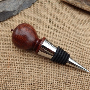 K&ouml;nnte beinhalten: Ein Weinflaschenverschluss aus Holz mit dunkelroter Oberfl&auml;che und einem silbernen Metallfu&szlig;. Der Verschluss hat eine Gummidichtung, um den Wein frisch zu halten.