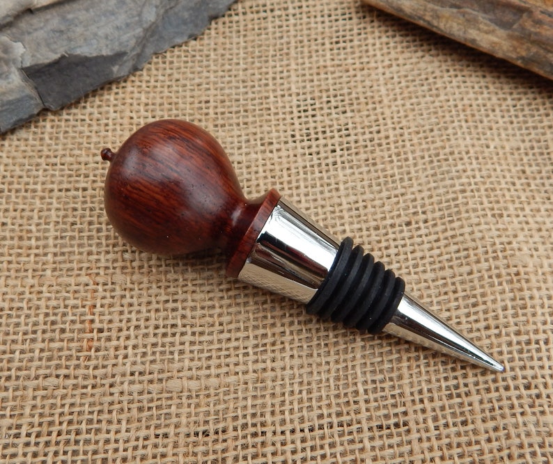 K&ouml;nnte beinhalten: Ein Weinflaschenverschluss aus Holz mit dunkelroter Oberfl&auml;che und einem silbernen Metallfu&szlig;. Der Verschluss hat eine Gummidichtung, um den Wein frisch zu halten.
