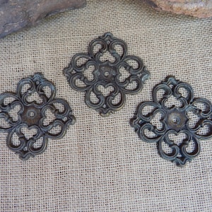 May include: Three dark bronze-colored metal decorative rosettes featuring an intricate openwork scroll pattern. Each square-shaped piece has a central circular hole and symmetrical, curved filigree details radiating outward from the center point.