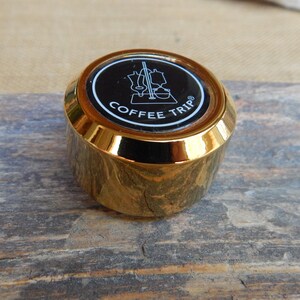 May include: Gold-colored metal bottle stopper with a black circle featuring a white outline of a coffee maker and the text "COFFEE TRIP" in white.
