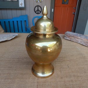 May include: A gold-colored, decorative urn with a lid. The urn has a bulbous body, a pedestal base, and a domed lid with a pointed finial. The surface of the urn is highly polished, reflecting the surrounding environment.