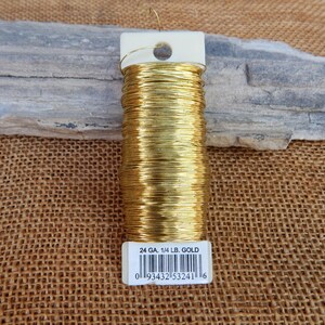 May include: A spool of gold-colored floral wire, labeled "24 GA. 1/4 LB. GOLD." The wire is tightly wound around a white plastic spool. The background includes a piece of gray stone and a burlap surface.