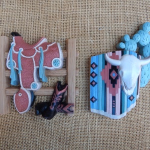 May include: Two decorative wall hangings with a western theme. The first features a saddle and boots hanging on a wooden fence. The second features a cow skull with a turquoise and brown patterned blanket behind it.
