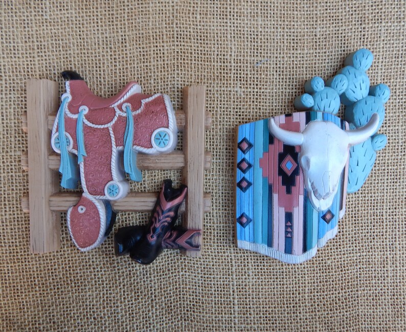 May include: Two decorative wall hangings with a western theme. The first features a saddle and boots hanging on a wooden fence. The second features a cow skull with a turquoise and brown patterned blanket behind it.