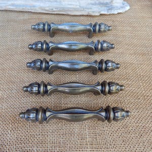 May include: Five antique bronze cabinet pulls with ornate detailing. Each pull features a curved handle and decorative end pieces. The pulls are arranged on a textured, neutral-toned surface, showcasing their vintage aesthetic.