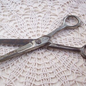 May include: Vintage silver-toned barber shears with a rounded handle and a single finger ring. The shears are lying on a white crocheted doily.