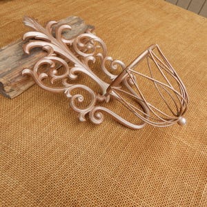 May include: A decorative wall sconce in a metallic rose gold finish. The sconce features an ornate, scrolling design with a basket-like holder. The design includes a series of swirling patterns and a rounded base, perfect for displaying small items.