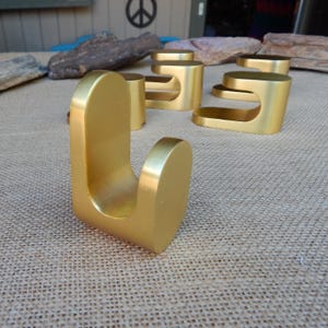 May include: A collection of gold-colored metal hooks, with a curved design. The hooks are arranged on a textured surface, with a peace sign visible in the background. The hooks are designed for hanging items.