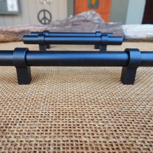 May include: Three black metal cabinet pulls with a cylindrical shape and square ends. The pulls are shown on a burlap surface.