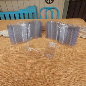 May include: Clear plastic hinged boxes, stacked in groups, tied with white string. Two individual boxes are open, displaying the interior. The boxes are on a brown burlap surface, with a blue chair and other objects in the background.