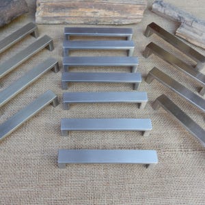 May include: A collection of fourteen rectangular cabinet pulls made of brushed metal with a matte silver finish. Each pull features a simple, straight bar design with two perpendicular mounting posts, arranged in three distinct groups on a surface.