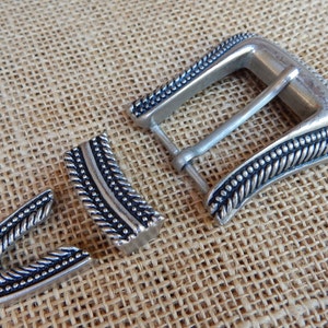 K&ouml;nnte beinhalten: Silberne G&uuml;rtelschnalle mit geflochtenem Design und schwarzen Akzenten. Die Schnalle besteht aus drei Teilen, der Hauptschnalle und zwei kleineren Teilen, die am G&uuml;rtel befestigt werden.