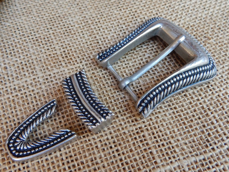 K&ouml;nnte beinhalten: Silberne G&uuml;rtelschnalle mit geflochtenem Design und schwarzen Akzenten. Die Schnalle besteht aus drei Teilen, der Hauptschnalle und zwei kleineren Teilen, die am G&uuml;rtel befestigt werden.