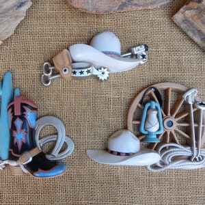May include: A set of five western-themed wall decor pieces. The set includes a cowboy hat, a boot, a cactus, a wagon wheel, and a lantern. The pieces are made of a light brown material and have a rustic look.