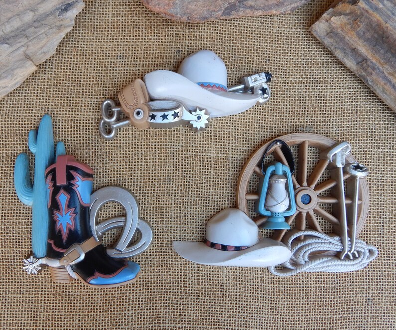 May include: A set of five western-themed wall decor pieces. The set includes a cowboy hat, a boot, a cactus, a wagon wheel, and a lantern. The pieces are made of a light brown material and have a rustic look.