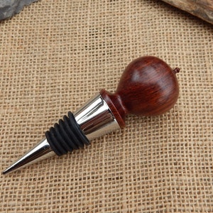 K&ouml;nnte beinhalten: Ein Weinflaschenverschluss aus Holz mit dunkelroter Oberfl&auml;che und einem silbernen Metallfu&szlig;. Der Verschluss hat eine Gummidichtung, um den Wein frisch zu halten.