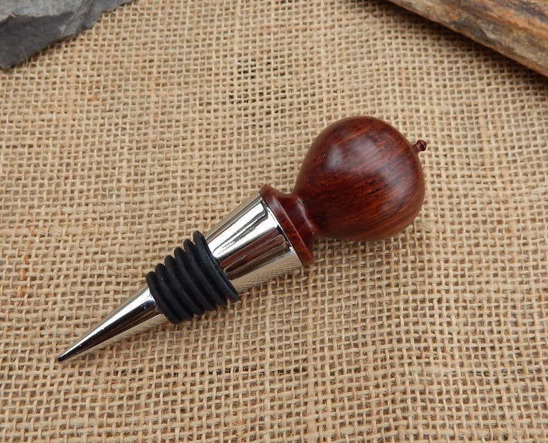 K&ouml;nnte beinhalten: Ein Weinflaschenverschluss aus Holz mit dunkelroter Oberfl&auml;che und einem silbernen Metallfu&szlig;. Der Verschluss hat eine Gummidichtung, um den Wein frisch zu halten.