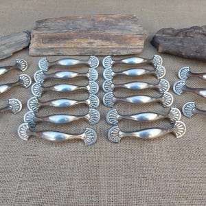 May include: A collection of silver-toned cabinet pulls with a decorative fan-shaped design on each end. The pulls are arranged on a neutral-colored burlap surface, with pieces of wood in the background.