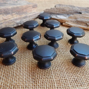 May include: Twelve black octagonal cabinet knobs with a brushed bronze finish. The knobs are arranged in a circle on a burlap surface.