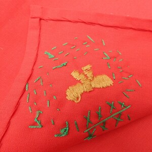 May include: A folded red fabric with embroidered details. The embroidery features a tan figure and green stitches scattered around it. The fabric is a solid, vibrant red color.