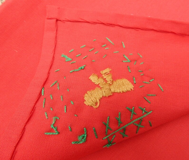 May include: A folded red fabric with embroidered details. The embroidery features a tan figure and green stitches scattered around it. The fabric is a solid, vibrant red color.