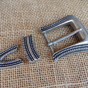K&ouml;nnte beinhalten: Eine silberne G&uuml;rtelschnalle mit einem dekorativen Seil-Design und schwarzen Akzenten. Die Schnalle ist in drei Teile unterteilt, darunter die Hauptschnalle, eine Zunge und ein Halter.