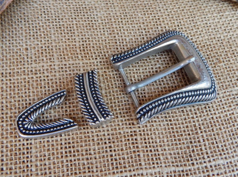 K&ouml;nnte beinhalten: Eine silberne G&uuml;rtelschnalle mit einem dekorativen Seil-Design und schwarzen Akzenten. Die Schnalle ist in drei Teile unterteilt, darunter die Hauptschnalle, eine Zunge und ein Halter.