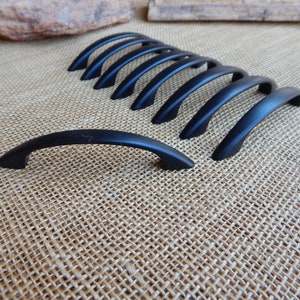 May include: A set of ten black metal cabinet pulls with a curved design. The pulls are arranged in a row on a burlap surface.