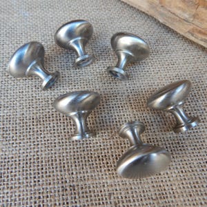 May include: Six brushed nickel cabinet knobs with an oval top and a short, rounded stem. The knobs are arranged on a textured, tan surface, suggesting a rustic or vintage aesthetic. The hardware is designed for furniture or cabinetry.