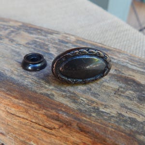 May include: An antique-style oval cabinet knob with a dark finish and decorative border. A matching small, round base is next to the knob. The knob and base are on a weathered wooden surface.