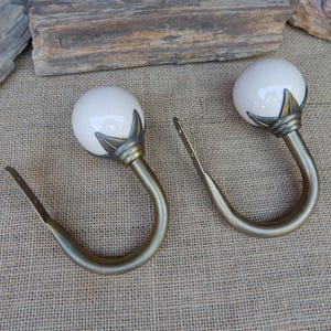 May include: Two curtain tiebacks featuring a smooth, round, off-white ceramic ball finial attached to a curved, antique brass-toned metal hook. Each hook has a decorative leaf-shaped base where it connects to the ceramic sphere and a flat mounting plate.