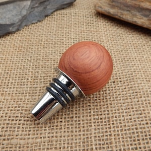 May include: A wooden bottle stopper with a round, reddish-brown top and a silver base. The stopper has a black rubber seal.