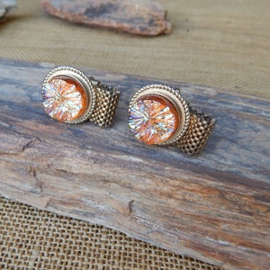 May include: A pair of vintage cufflinks with iridescent orange centers and gold-tone metal settings. The round centers have a textured, starburst-like appearance. The cufflinks have a woven metal band.