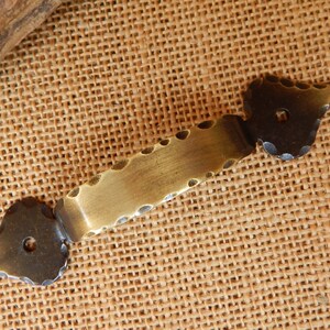 May include: A brass drawer pull with a scalloped edge and two black heart-shaped ends. The pull has two screw holes.