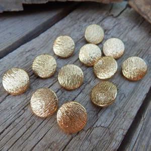 May include: A collection of 11 gold-colored buttons with a textured surface. The buttons are round and have a slightly raised edge.