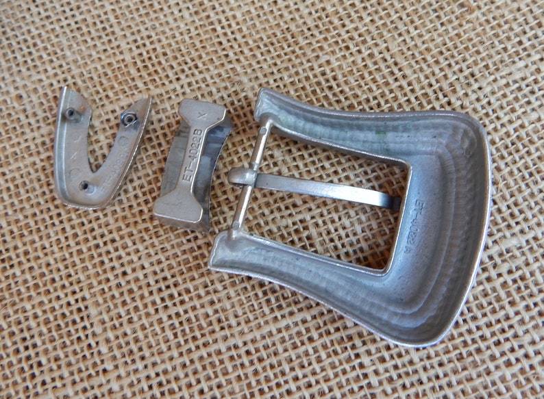 K&ouml;nnte beinhalten: Eine silberne G&uuml;rtelschnalle aus Metall mit dem Text "ET-1022B" auf der R&uuml;ckseite. Die Schnalle besteht aus drei Teilen, der Hauptschnalle, einem kleineren Teil mit dem Text und einem separaten Teil, der den G&uuml;rtel h&auml;lt.