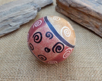 May include: A round, hand-painted ceramic sphere with a geometric design. The sphere is decorated with swirls in black, white, and red on a tan background.