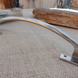 May include: A silver-toned, curved cabinet pull with a rectangular base. The pull is shown on a textured, beige surface.