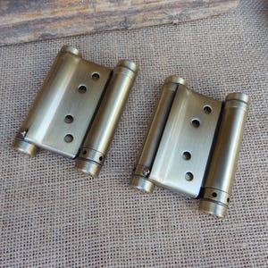 May include: Two antique brass-colored double-action spring hinges. Each hinge has a rectangular plate with three circular holes and two cylindrical barrels. The hinges are designed to swing doors open in both directions.