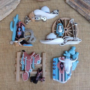 May include: A set of five western-themed wall decor pieces. The set includes a cowboy boot, a saddle, a cactus, a wagon wheel, and a skull with a turquoise and brown patterned blanket. The pieces are made of a light brown material and have a rustic, vintage look.