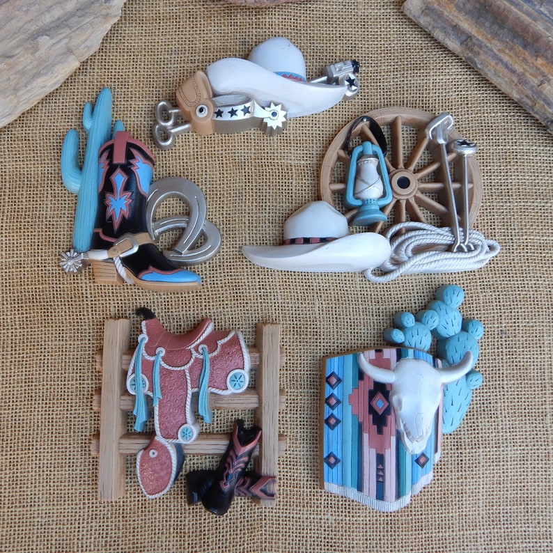 May include: A set of five western-themed wall decor pieces. The set includes a cowboy boot, a saddle, a cactus, a wagon wheel, and a skull with a turquoise and brown patterned blanket. The pieces are made of a light brown material and have a rustic, vintage look.