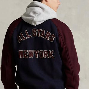 Puede incluir: Chaqueta universitaria azul marino y burdeos con el texto "ALL STARS NEWYORK" en la espalda. La chaqueta tiene una capucha gris y una gorra azul marino con detalles dorados.