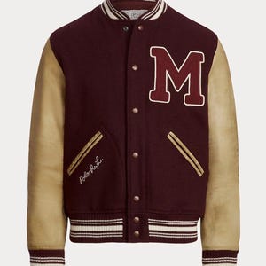 May include: Maroon varsity jacket with tan leather sleeves. Features a large white-outlined "M" patch, gold accents, and the embroidered text "Polo R.L.". The jacket has a striped collar, cuffs, and hem.