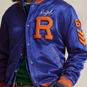 May include: A royal blue satin bomber jacket with orange accents on the collar, cuffs, and pocket trim. Features a large orange letter "R" and the name "Ralph" embroidered in white. The number "61" and chevron details are on the sleeve.