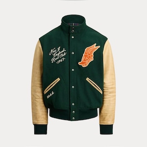 Op de afbeelding: Een groene en beige varsity jas met een hoge kraag en drukknoopsluiting. De jas heeft beige leren mouwen, een groene body en geborduurde patches met tekst en een gevleugeld voetlogo. De tekst luidt "New England Run Club - 1967."