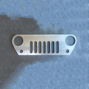 May include: A silver-colored metal Jeep-style grille with a series of vertical slots and two circular holes. The grille is designed to be mounted on a vehicle. The metal has a brushed finish.