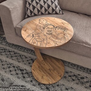 May include: A round wooden side table with a single curved support leg and a circular base. The tabletop features a dark, laser-engraved line art illustration of a person with glasses carrying two children, one on their shoulders and one beside.