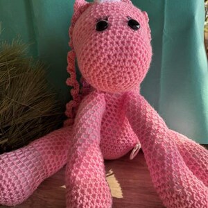 May include: A handmade pink crochet unicorn doll with a white horn and black eyes. The unicorn has long legs and a curly tail. The doll is made of soft yarn and is a whimsical toy.