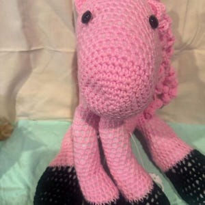 May include: A handmade pink crochet unicorn stuffed animal with black hooves and a striped horn. The unicorn has black button eyes and a pink mane. The toy is made of soft yarn and is suitable for children.